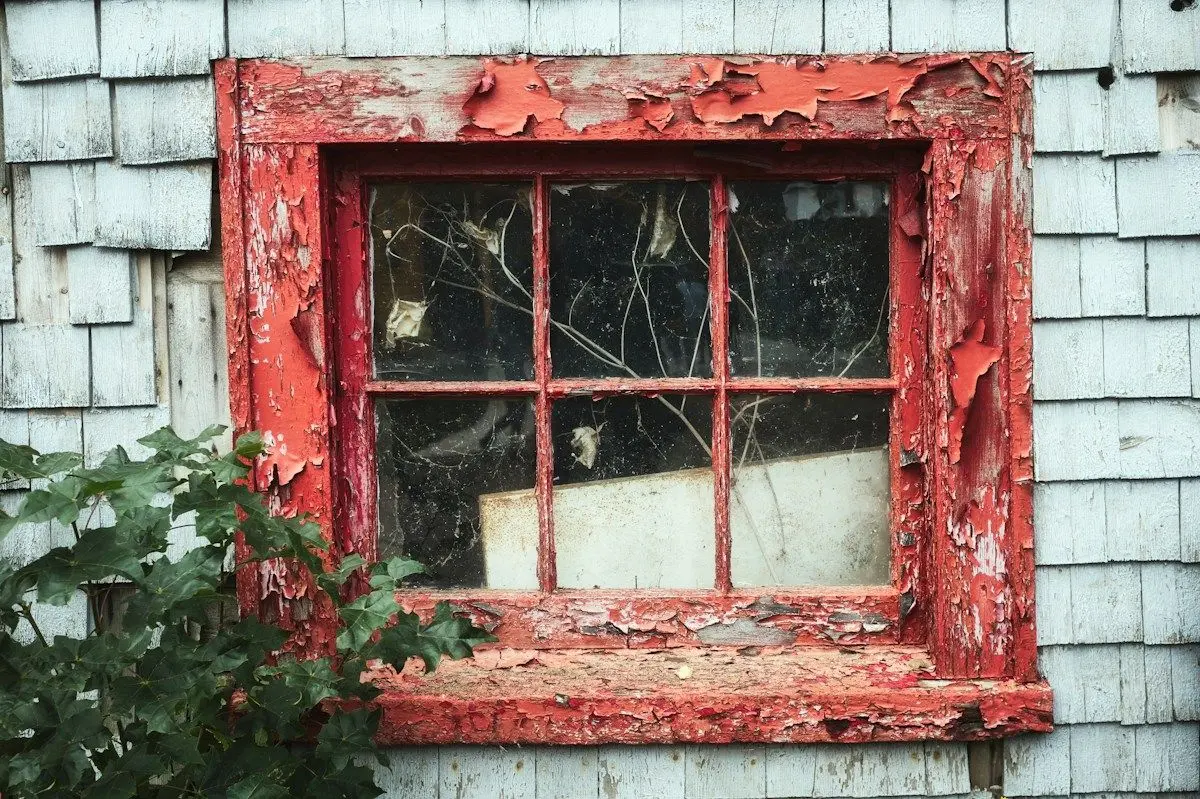 Old wood-frame window with peeling red paint on a shingled wall